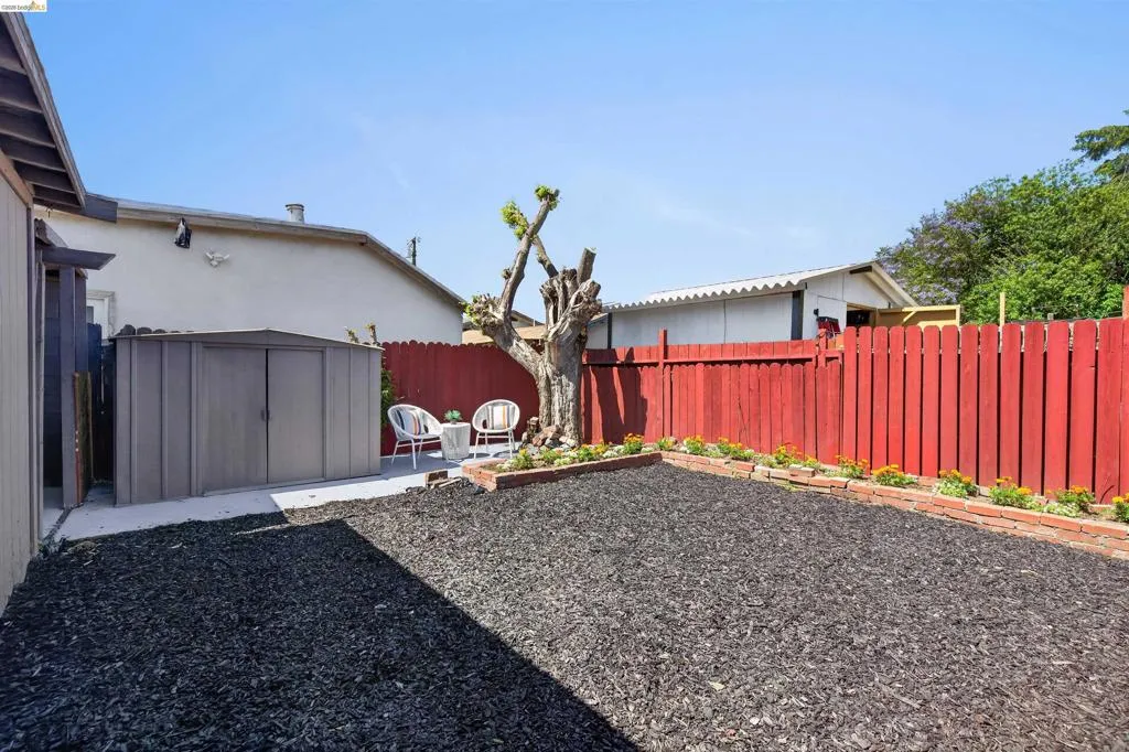 Rear yard featuring black mulch ground cover, a red wood fence, and a concrete patio area with an included storage shed