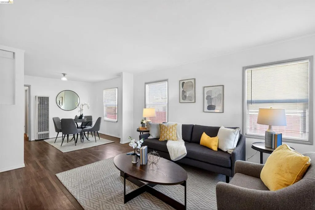 Open concept living and dining area featuring wood-finish flooring, white walls, multiple windows with blinds, and a built-in wall heater