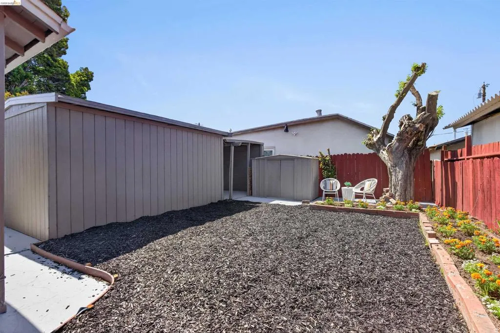 Spacious backyard featuring dark ground covering, a garden bed with blooming flowers, a mature tree, and red fencing