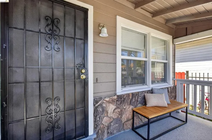 Front porch featuring a black iron security door with decorative scrollwork, tan siding, stone veneer accents, white-framed windows, and a white porch railing