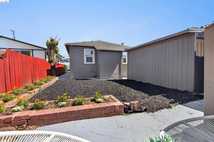 Backyard area featuring a gray exterior building, red perimeter fencing, and a brick-edged planting bed with small flowers