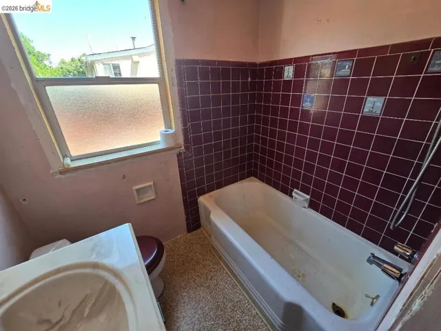 Bathroom featuring a full bathtub with tiled surround, a window providing natural light, and a toilet