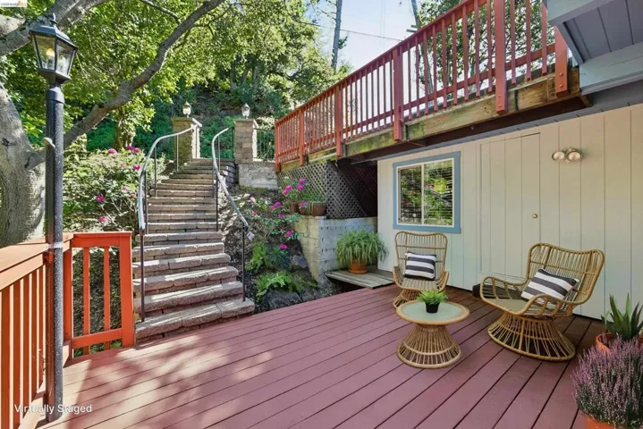 Expansive reddish-brown deck featuring an overhead balcony, a white paneled exterior wall with a light blue trimmed window, and a stone staircase with metal railings