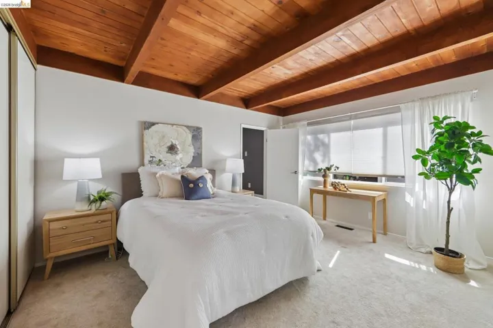 Vaulted wood-finish ceiling with exposed beams, light-toned wall finishes, carpeted flooring, and a window with white roller blinds and sheer curtains
