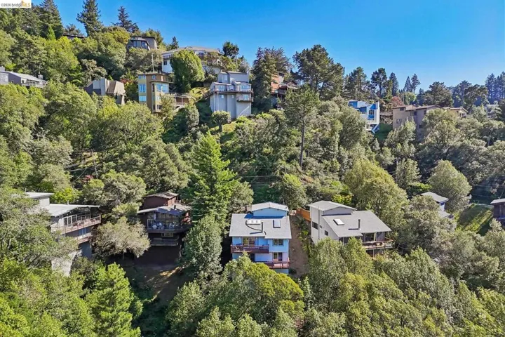 Hillside property surrounded by mature trees, featuring a light blue exterior, multi-level construction, and a private balcony