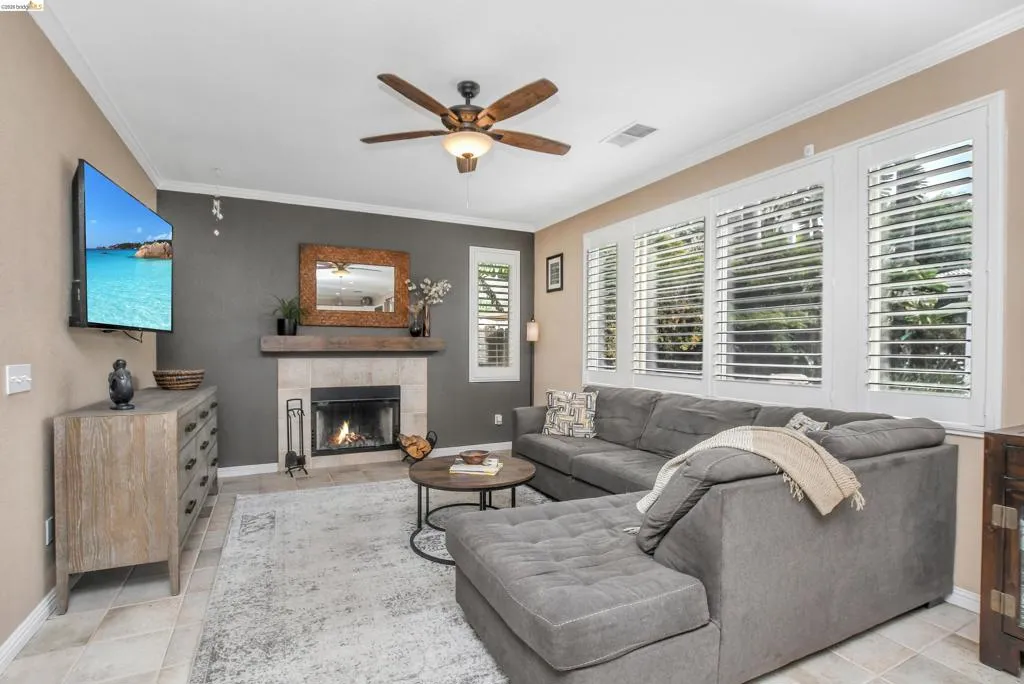 Living area featuring a tiled fireplace with a wood mantle, a ceiling fan with integrated lighting, multiple windows with white plantation shutters, and crown molding