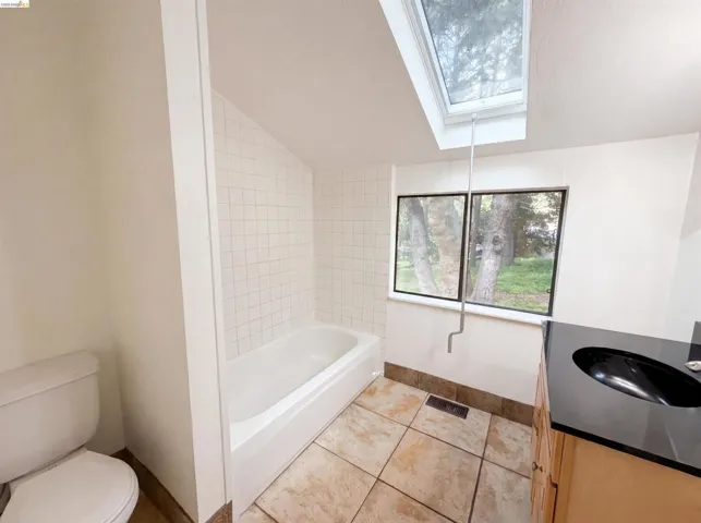Bathroom featuring a skylight, white subway tile bath surround, a white built-in tub, a wood-finish vanity with a black countertop and integrated sink, and large format floor tiling
