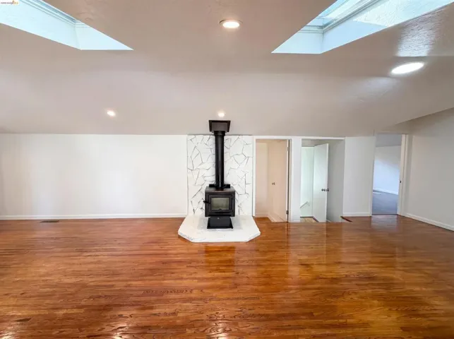 Spacious room featuring rich wood flooring, a central wood-burning stove with a stone-clad surround, and two ceiling skylights