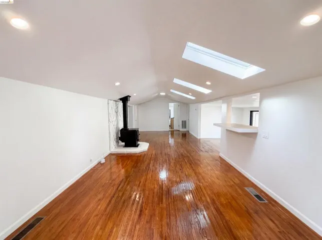 Expansive living area featuring warm wood-finish flooring, a vaulted ceiling with multiple skylights, recessed lighting, and a prominent wood-burning stove with a textured surround