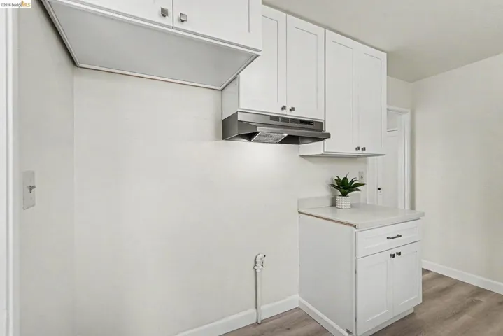 Kitchen area featuring white upper and lower cabinetry with silver-tone hardware, a stainless steel range hood, light-toned countertops, and wood-finish flooring