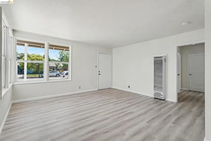 Spacious room featuring light wood-finish flooring, multiple windows with white trim, and a wall-mounted heating unit