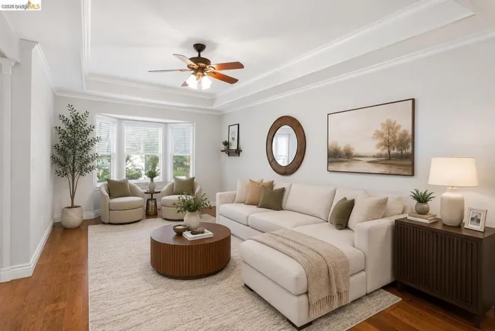 Living area featuring wood-finish flooring, a bay window with natural light, a tray ceiling with crown molding, and a ceiling fan AI photo