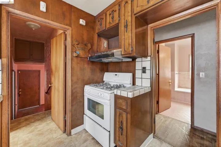 Kitchen featuring wood-paneled walls, wood cabinetry with decorative hardware, a white gas range with an overhead vent, and a tiled countertop