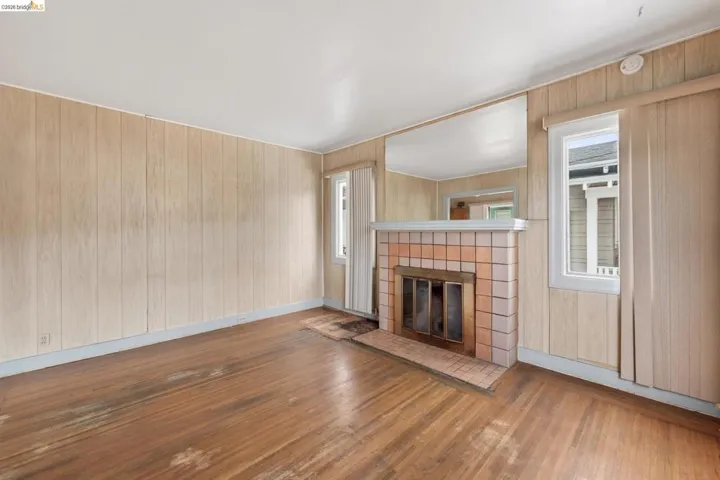 Room featuring wood-finish flooring, light-toned wood paneling, a tiled fireplace with a metal grate, and two windows with vertical blinds