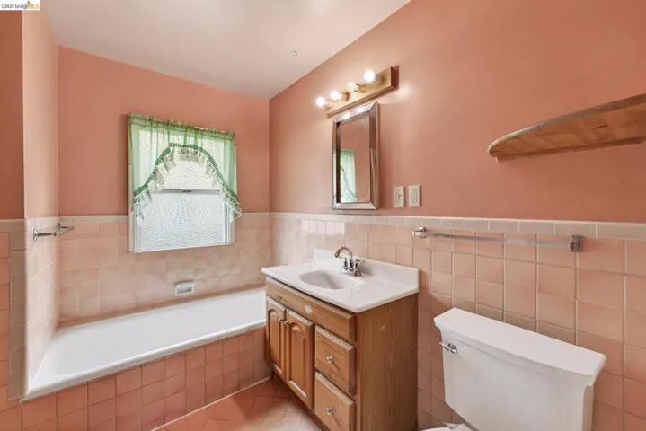 Bathroom featuring a built-in bathtub, a window with textured glass, a wood-finish vanity with a white countertop, a mirror with an overhead light fixture, and ceramic tile wainscoting