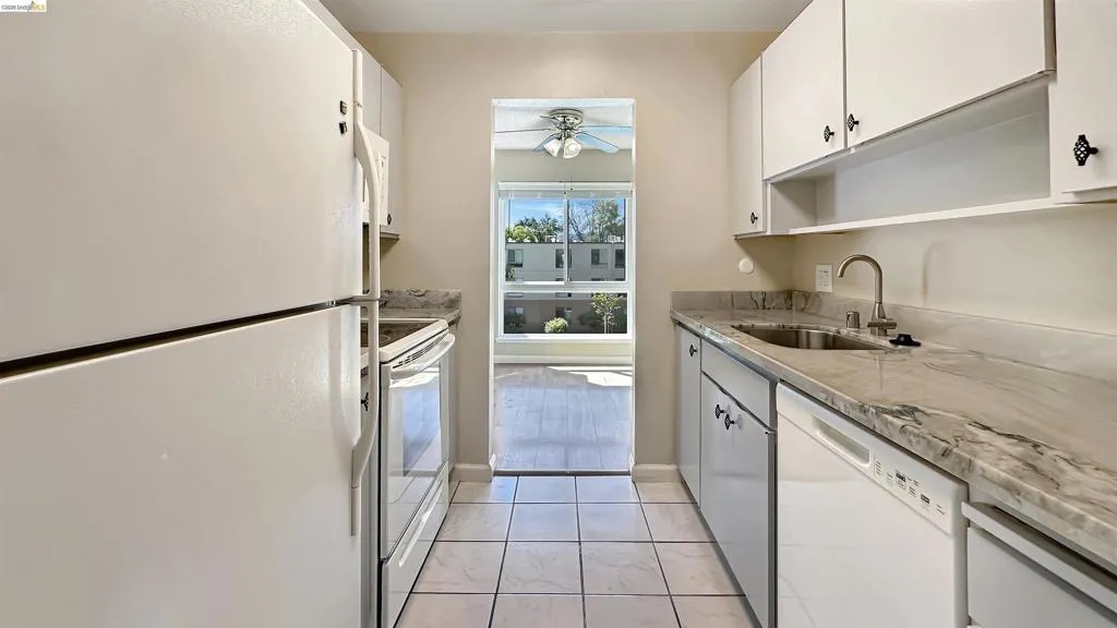 Galley kitchen featuring white cabinetry with decorative hardware, white appliances, patterned stone countertops, and tile flooring