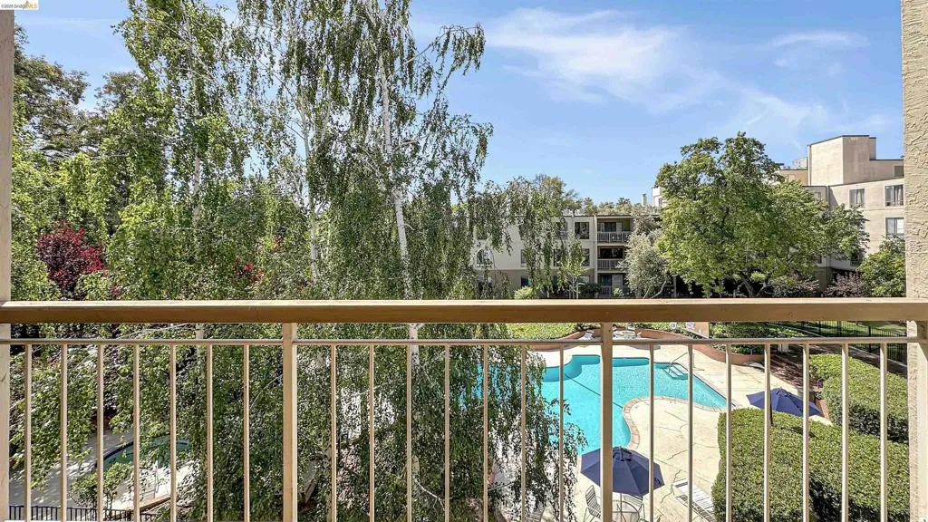 Balcony with metal railing overlooking a swimming pool, surrounding patio, lush green landscaping, and distant residential structures