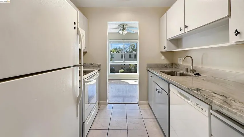 Galley kitchen featuring white cabinetry with decorative hardware, white appliances, patterned stone countertops, and tile flooring