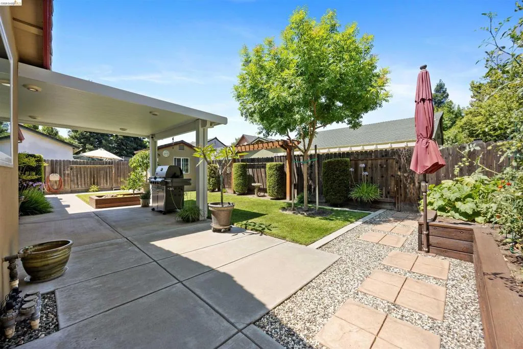 Covered concrete patio with recessed lighting, gravel and paver pathway, raised garden beds, wood fence perimeter, and established landscaping