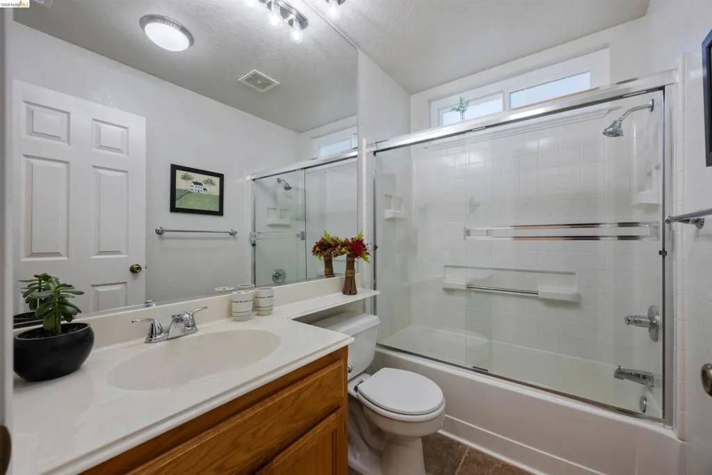 Bathroom featuring a wood-finish vanity with an integrated sink and chrome faucet, a shower-over-tub combination with sliding glass doors and white tile surround, and an overhead window providing natural light