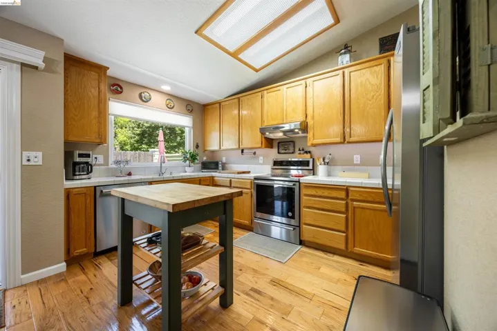 Kitchen featuring wood-finish flooring, natural wood cabinetry, white tile countertops, stainless steel appliances, and an overhead fluorescent light panel