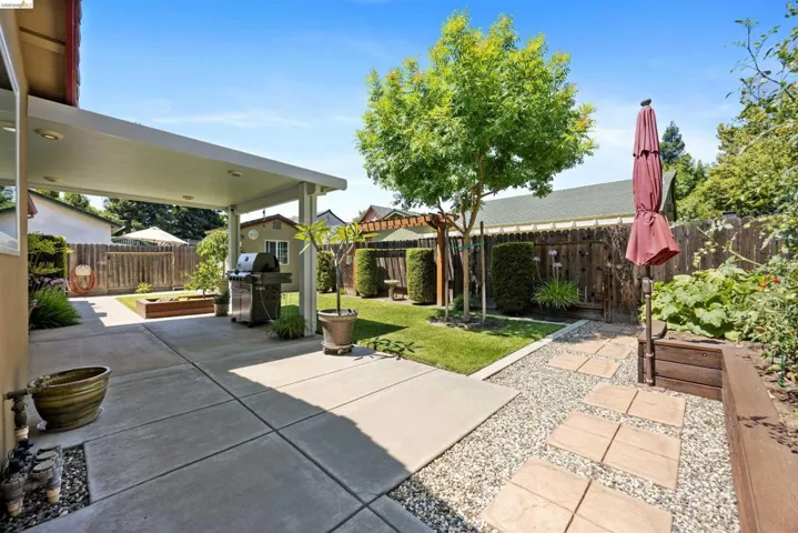 Covered concrete patio with recessed lighting, gravel and paver pathway, raised garden beds, wood fence perimeter, and established landscaping