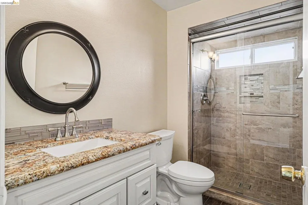 Bathroom featuring a white vanity with a granite-style countertop, a circular framed mirror, and a glass-enclosed shower with tiled walls and a mosaic accent