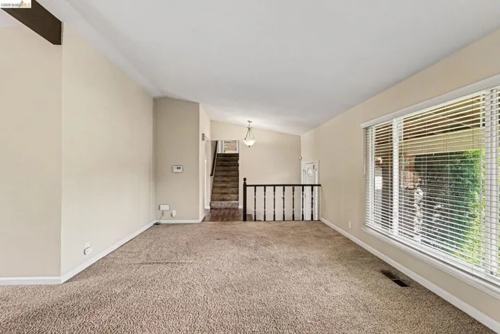 Spacious living area featuring neutral carpeting, a large window with white blinds, and a vaulted ceiling with a visible beam