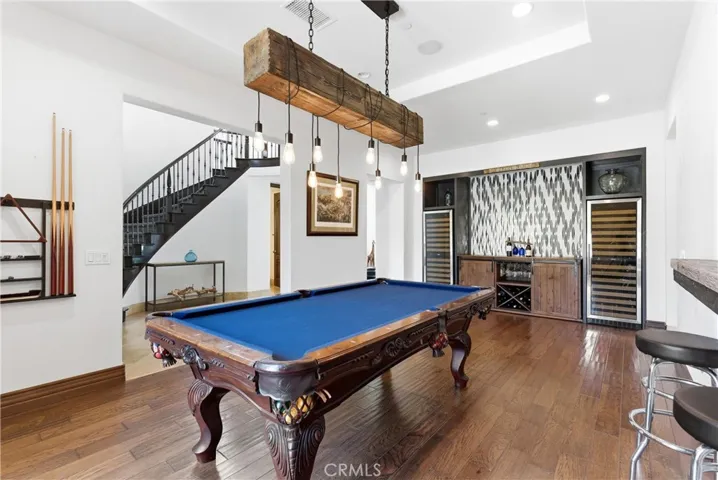 Entertainer’s game room with custom billiards area, rustic beam chandelier, built-in wet bar, dual wine refrigerators, hardwood flooring, and open-concept design.