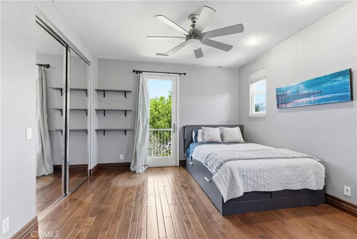 Bright upstairs guest bedroom with hardwood flooring, mirrored closet doors, built-in shelving, and private balcony access overlooking mature greenery.