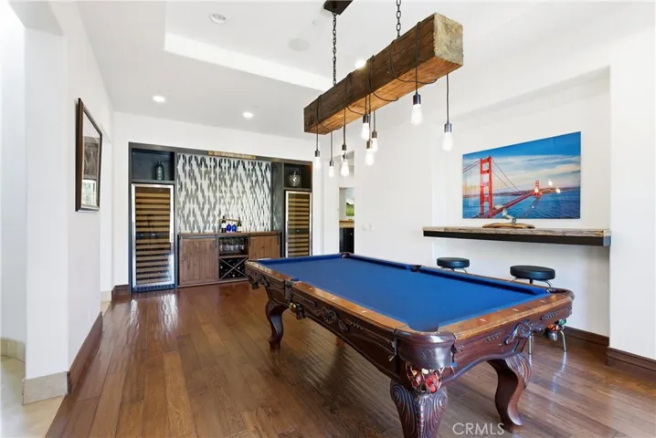 Entertainer’s game room with billiards area, custom wet bar, dual wine refrigerators, hardwood flooring, designer lighting, and built-in beverage station.