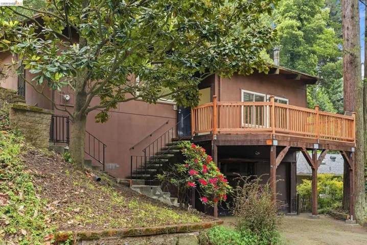 Terraced exterior featuring a spacious elevated deck with wood railings, a dedicated stairway entry, natural wood-finish siding, and mature landscaping
