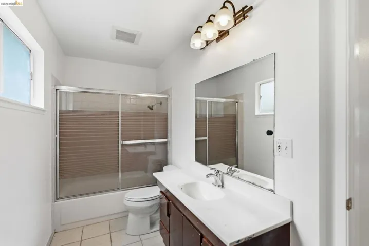 Bathroom featuring a white tile floor, a shower-tub combination with sliding glass doors, a vanity with a white countertop and integrated sink, a large wall-mounted mirror, and overhead lighting