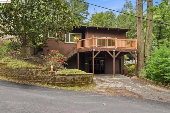 Brown exterior with a ground-level driveway, elevated wooden deck, and mature surrounding trees