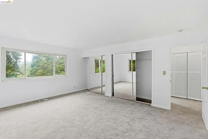 White-painted room featuring light gray carpeting, a wide window with a tree-filled outdoor scene, and two sets of built-in closet doors, one mirrored and one louvered