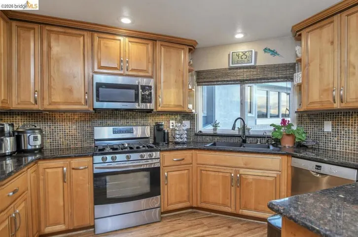 Kitchen with stainless steel appliances, brown cabinets, dark stone countertops, light wood-style floors, and tasteful backsplash