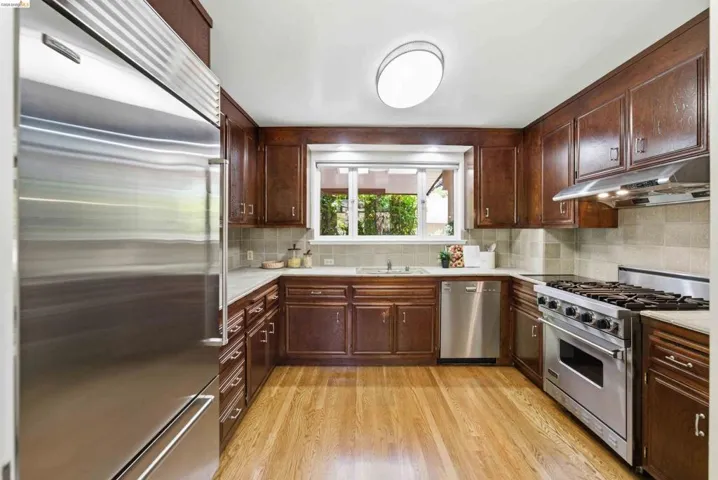 Kitchen featuring wood-finish flooring, dark wood cabinetry, a built-in stainless steel refrigerator, stainless steel dishwasher, and a stainless steel range with a ventilation hood