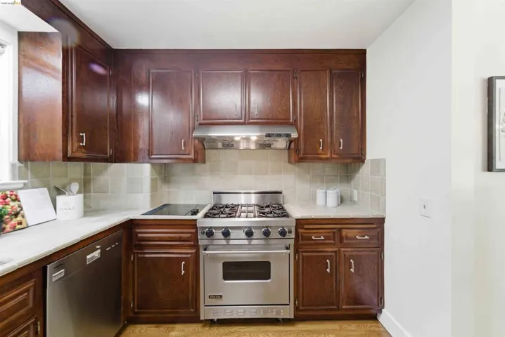 Kitchen featuring wood cabinetry, light-toned countertops, tile backsplash, stainless steel gas range, and a built-in dishwasher
