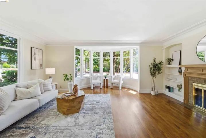Spacious interior featuring a multi-pane bay window, wood-finish flooring, built-in shelving, a fireplace with a wood mantle, and crown molding
