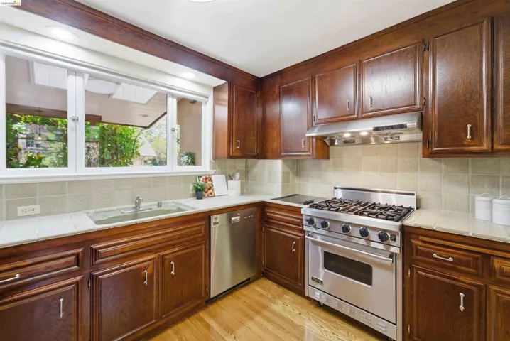 Kitchen featuring rich wood cabinetry, tile backsplash, white countertops, stainless steel appliances, and a window overlooking exterior landscaping