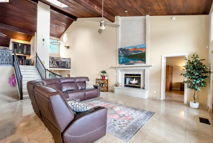 Grand living area featuring a vaulted wood-finish ceiling, a stone fireplace with a hearth, polished tile flooring, a two-story staircase with ornate iron railings, and a skylight
