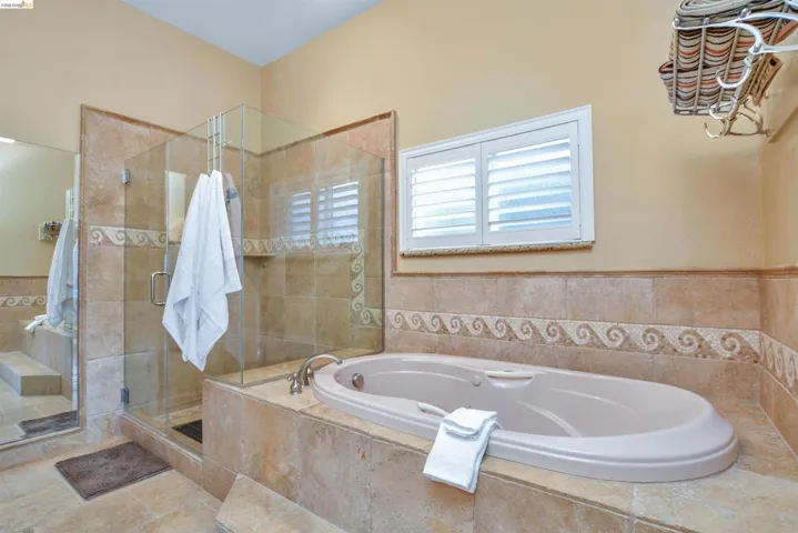 Bathroom featuring a built-in soaking tub, glass-enclosed shower, stone-finish tile work, and a window with plantation shutters