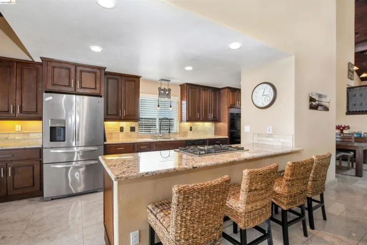 Spacious kitchen featuring an expansive granite-top island with a built-in gas cooktop, rich wood cabinetry, stainless steel appliances, and a mosaic tile backsplash