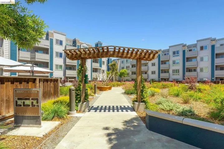Path with an overhead wood pergola, bordered by landscaped plantings, leading towards multi-story residential buildings with balconies
