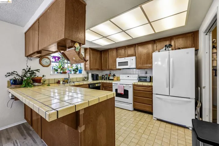 Kitchen featuring wood cabinetry, white appliances, fluorescent ceiling light, tile flooring, and a tile countertop peninsula
