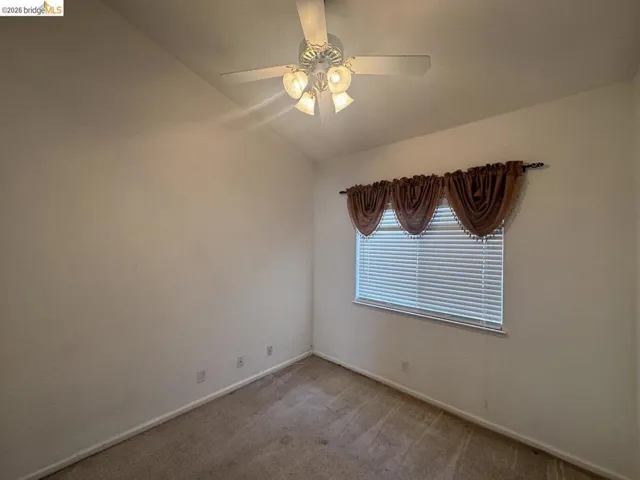 Room featuring a prominent window with horizontal blinds and valance, overhead ceiling fan with light fixture, vaulted ceiling, and neutral-toned carpeting