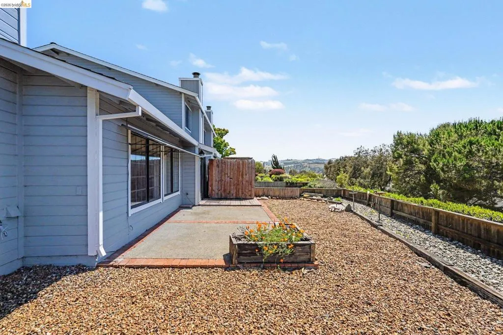 Rear exterior featuring a concrete patio with brick edging, a gravel landscape, a wooden fence, and multiple windows