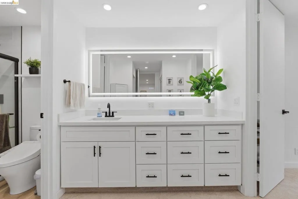 Dual-sink vanity with white cabinetry and dark hardware, quartz countertop, and an illuminated mirror