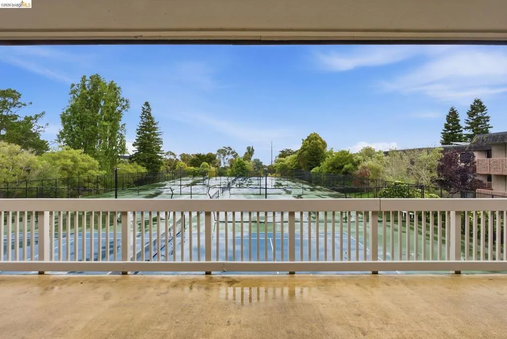 Balcony with concrete flooring and a white railing overlooking multiple tennis courts, surrounded by mature trees and lush landscaping