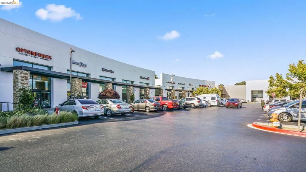 Modern commercial center with a spacious asphalt parking lot, contemporary storefronts featuring large glass windows, stone veneer columns, and dark awnings